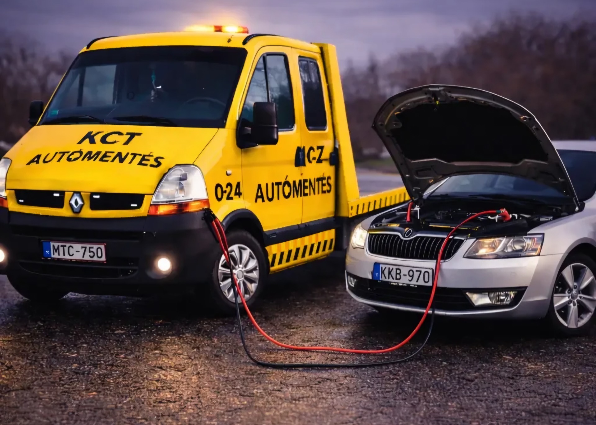 Sárga mentőautó és fehér autó nyitott motorháztetővel, piros kábelekkel, szürkületben, úton.